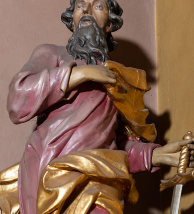 Zdjęcie nr 3: Rzeźba całopostaciowa przedstawia stojącego w wydatnym kontrapoście, na niewysokim postumencie św. Pawła zwróconego na wprost, z głową skierowaną w trzech czwartych w prawo. Twarz świętego charakteryzuje śniada karnacja, wyraziste rysy oraz długi, brązowy zarost. Półdługie, kręcone włosy opadają na kark mężczyzny. Święty ma na sobie długą, jasnoliliową suknię. Połę narzuconego na lewe ramię, rozwianego, złoconego płaszcza osłaniającego lewe biodro, podtrzymuje na wysokości klatki piersiowej prawą ręką. W lewej, zgiętej w łokciu i uniesionej na wysokości talii podtrzymuje miecz o złoconej rękojeści i srebrzonym ostrzu skierowanym w dół. 