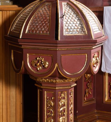 Zdjęcie nr 3: Płaskorzeźba w kształcie stojącego prostokąta zamkniętego łukiem półkolistym przedstawia scenę Chrztu Jezusa w Jordanie. Na pierwszym planie, po prawej stronie kompozycji znajduje się stojąca w lekkim kontrapoście postać Jezusa zwróconego nieznacznie w lewo. Jego postawę charakteryzuje lekko pochylona głowa z krótkim zarostem, o długich, falowanych włosach oraz skrzyżowane na piersiach ręce. Nagą sylwetkę osłania jedynie w partii bioder, sięgające połowy ud białe perizonium. Jezus stoi w płynącej środkiem kompozycji rzece. Po lewej stronie, na skalistym brzegu stoi zwrócony w trzech czwartych w prawo św. Jan Chrzciciel. Lekko pochyloną głowę charakteryzuje krótki ciemnobrązowy zarost i długie, opadające na plecy włosy. Nagą sylwetkę mężczyzny osłania w partii bioder perizonium wykonane ze skóry zwierzęcej. Prawą ręką podtrzymuje długą laskę w formie krzyża z banderolą z napisem: „Ecce Agnus Dei”, lewą, wyciągniętą nad głową Jezusa wylewa z małej, płaskiej miseczki wodę. Tło przedstawienia stanowi rozległy pejzaż z wzniesieniami, z widoczną na drugim planie murowaną zabudową oraz smukłymi drzewami palmowymi w głębi. Błękitne niebo zaczerwienione jest tuż nad linią horyzontu. 
Płaskorzeźbę ujmuje rzeźbiona na wzór retabulum ołtarzowego architektoniczna struktura. Podstawę tworzy wysoki cokół, ujęty parą kolumienek, którego pole wypełnia płycina w kształcie stojącego prostokąta o wklęsło ściętych narożach. Na cokole osadzona jest część środkowa struktury, w formie prostokątnej arkady zamkniętej łukiem półkolistym, ujętej parą kolumienek oraz parą uszaków w postaci zamkniętych trójkątnie przypór. Struktura powyżej arkady przybiera formę frontonu budowli ze szczytem, jaki tworzy gzyms wieńczący w kształcie trójkątnego przyczółka, flankowany parą niewielkich pilastrów. Powyżej widoczna konstrukcja przypominająca pokryty dachówką dach pulpitowy. Zwieńczenie ma formę aediculi z arkadą z łukiem trójlistnym, ujętą parą kolumienek wspierających belkowanie z gzymsem w kształcie półkolistego łuku nadwieszonego. Dodatkowo po bokach aediculi znajduje się para przypór. Powyżej arkady w części środkowej przedstawiono płaskorzeźbione  popiersie Boga Ojca. W arkadzie zwieńczenia jest figura anioła. Utrzymaną w odcieniach brązu strukturę zdobi złocona dekoracja ornamentalna w formie stylizowanych wici roślinnych i rozet.       

