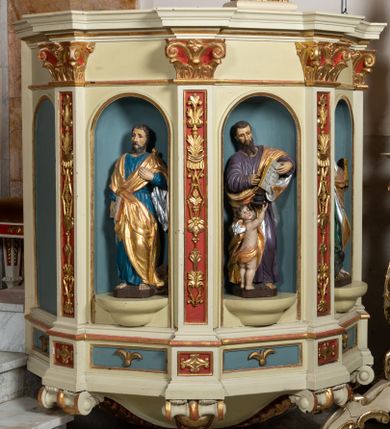 Zdjęcie nr 1: Kosz ambony wykonano na rzucie ośmioboku, wspartego na ośmiobocznym cokole. Bok kosza artykułują pilastry o kompozytowych głowicach i dekorowanych arabeską trzonach, wspartych na postumentach ozdobionych ornamentem geometrycznym. Pilastry podtrzymują profilowane belkowanie wyłamane na ich osi. Pomiędzy pilastrami umieszczono półokrągłe nisze zaopatrzone u spodu w półokrągłe konsole. W niszach ustawiono całopostaciowe, rzeźbione przedstawienia czterech Ewangelistów. Pod amboną podwieszono wolutowe spływy łączące cokół kolejnego pilastru ze znajdującą się na spodzie podwieszenia dekoracyjną szyszką. Na belkowaniu na osi usytuowano pulpit w formie rozwiniętego zwoju pisarskiego wspartego na powiększonym kapitelu kompozytowym, wzorowanym na kapitelu z pilastrów bocznych. Strukturę architektoniczną kosza polichromowanego w kolorze ciemnej kości słoniowej, wnętrze wnęk na niebiesko, a tło trzonów kapiteli i ich głowic na czerwono. Rzeźby czterech Ewangelistów uzyskały polichromię naturalistyczną. 