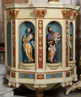 Zdjęcie nr 1: Kosz ambony wykonano na rzucie ośmioboku, wspartego na ośmiobocznym cokole. Bok kosza artykułują pilastry o kompozytowych głowicach i dekorowanych arabeską trzonach, wspartych na postumentach ozdobionych ornamentem geometrycznym. Pilastry podtrzymują profilowane belkowanie wyłamane na ich osi. Pomiędzy pilastrami umieszczono półokrągłe nisze zaopatrzone u spodu w półokrągłe konsole. W niszach ustawiono całopostaciowe, rzeźbione przedstawienia czterech Ewangelistów. Pod amboną podwieszono wolutowe spływy łączące cokół kolejnego pilastru ze znajdującą się na spodzie podwieszenia dekoracyjną szyszką. Na belkowaniu na osi usytuowano pulpit w formie rozwiniętego zwoju pisarskiego wspartego na powiększonym kapitelu kompozytowym, wzorowanym na kapitelu z pilastrów bocznych. Strukturę architektoniczną kosza polichromowanego w kolorze ciemnej kości słoniowej, wnętrze wnęk na niebiesko, a tło trzonów kapiteli i ich głowic na czerwono. Rzeźby czterech Ewangelistów uzyskały polichromię naturalistyczną. 