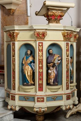 Zdjęcie nr 1: Kosz ambony wykonano na rzucie ośmioboku, wspartego na ośmiobocznym cokole. Bok kosza artykułują pilastry o kompozytowych głowicach i dekorowanych arabeską trzonach, wspartych na postumentach ozdobionych ornamentem geometrycznym. Pilastry podtrzymują profilowane belkowanie wyłamane na ich osi. Pomiędzy pilastrami umieszczono półokrągłe nisze zaopatrzone u spodu w półokrągłe konsole. W niszach ustawiono całopostaciowe, rzeźbione przedstawienia czterech Ewangelistów. Pod amboną podwieszono wolutowe spływy łączące cokół kolejnego pilastru ze znajdującą się na spodzie podwieszenia dekoracyjną szyszką. Na belkowaniu na osi usytuowano pulpit w formie rozwiniętego zwoju pisarskiego wspartego na powiększonym kapitelu kompozytowym, wzorowanym na kapitelu z pilastrów bocznych. Strukturę architektoniczną kosza polichromowanego w kolorze ciemnej kości słoniowej, wnętrze wnęk na niebiesko, a tło trzonów kapiteli i ich głowic na czerwono. Rzeźby czterech Ewangelistów uzyskały polichromię naturalistyczną. 