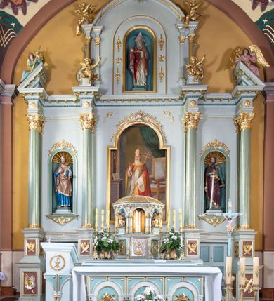 Zdjęcie nr 1: Nastawa ołtarza głównego przyjęła formę przyściennej, trójosiowej, jednokondygnacyjnej struktury architektonicznej ze wieńczeniem. Drewniany stipes ołtarzowy uzyskał kształt leżącego prostopadłościanu podtrzymującego mensę z portatylem, ustawionego na jednostopniowym podeście. Front stipesu udekorowano trzema płycinami obwiedzionymi profilowanymi listwami z rzeźbionymi ornamentami w polach. Na mensie ustawiono drewniane tabernakulum na rzucie trapezu. W dolnej części artykułują je czworoboczne płyciny z ornamentem geometrycznym, rozdzielone spływami wolutowymi podtrzymującymi profilowany gzyms, a w górnej części rozbudowany tron wystawienia. Prostopadłościenne, pancerne tabernakulum usytuowano na osi zdobiąc jego czworoboczne drzwiczki płyciną z trybowanym przedstawieniem kielicha w otoczeniu winnej latorośli i kłosów zbóż. Tron wystawienia przyjął formę trójarkadowej kapliczki zwieńczonej gzymsem koronującym i spłaszczonym dachem pokrytym złoconą rybią łuską. Arkady wsparto na korynckich, kanelowanych kolumienkach. W przyłuczach arkady środkowej, wypełnionej niszą na monstrancję, ukazano uskrzydlone dwie główki anielskie, a w przyłuczach arkad skrajnych motywy ornamentalne. Pod arkadami w osiach skrajnych umieszczono w niszach klęczących na jedno kolana na postumentach adorujących aniołów. Wsparty na arkadach gzyms koronujący ozdobiono profilowaniem i rytmicznie powtarzającymi się konsolkami. Za stipesem ustawiono strukturę ołtarzowa wspartą na wysokim cokole, artykułowaną czterema wysuniętym do przodu dwustopniowymi postumentami dekorowanymi profilowanym gzymsowaniem oraz prostokątnymi płycinami ujętymi w profilowane obramienia. W górnej strefie w płycinach od frontu wyobrażono uskrzydlone głowy symboli czterech Ewangelistów. Idę od lewej jest to głowa woła, anioła, orła i lwa. Natomiast w dolne strefie w płycinach znalazły się od lewej: skrzyżowane bicze z trzciną, na tle których wyobrażono chustę Weroniki, świecznik siedmioramienny z miedzianym wężem na palu na tle pektorału żydowskiego arcykapłana, tablice Dekalogu na tle skrzyżowanych trąb oraz krzyż skrzyżowany z kolumną biczowania wraz z lancą i gąbką na kiju. Pomiędzy postumentami w osiach skrajnych partii cokołowej usytuowano dwoje drzwi prowadzących do zakrystii, zdobionych czworobocznymi płycinami. Na postumentach ustawiono cztery kanelowane kolumny o kapitelach kompozytowych z impostami podtrzymujących belkowanie wyłamane na ich osi. Pomiędzy kolumnami artykułującymi kondygnację w osi środkowej umieszczono pole obrazowe, a w osiach skrajnych półkoliste nisze z ustawionymi w nich na postumentach figurami ŚŚ. Apostołów Piotra i Pawła. Zarówno nad polem obrazowym, jak i nad niszami oraz pod nimi widoczny jest ornament roślinny. Pole obrazowe na osi retabulum przyjęło kształt stojącego prostokąta z nadwieszonym półkolistym łukiem odcinkowym. Ujmuje je profilowana rama. W polu umieszczono obraz Wniebowzięcia Najświętszej Marii Panny, a na dwóch zasuwach przedstawienie św. Urbana oraz św. Barbary. Na belkowaniu w osiach skrajnych nad kolumnami zewnętrznymi usytuowano dwa przerwane przyczółki z leżącymi na nich parą aniołów trzymających krzyż i kotwicę. Natomiast na osi kolumn wewnętrznych na postumentach siedzą dwa złocone putta. Zwieńczenie ołtarza przyjęło kształt leżącego prostokąta flankowanego przez pilaster i kolumienkę podtrzymujących odcinki wyłamanego belkowania, na którym wsparto półkolisty, profilowany łuk odcinkowy wieńczący całość. Ponad tymi podporami umieszczono złocone, siedzące putta, a na osi łuku wieńczącego w promieniach z obłoczkami gołębicę Ducha Świętego. W polu zwieńczenia usytuowano półkolistą niszę ze stojącą figurą Boga Ojca. Ujmuje ją półkoliste obramienie zdobione stylizowaną ornamentyką roślinno-geometryczną. Strukturę architektoniczną retabulum polichromowano w tonacji jasnopastelowej zieleni, ornamenty, profile architektoniczne zostały wyzłocone, natomiast figury apostołów, Boga Ojca oraz aniołów przy tabernakulum i w zwieńczeniu w osiach skrajnych zostały naturalistycznie polichromowane. 