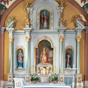 Zdjęcie nr 1: Nastawa ołtarza głównego przyjęła formę przyściennej, trójosiowej, jednokondygnacyjnej struktury architektonicznej ze wieńczeniem. Drewniany stipes ołtarzowy uzyskał kształt leżącego prostopadłościanu podtrzymującego mensę z portatylem, ustawionego na jednostopniowym podeście. Front stipesu udekorowano trzema płycinami obwiedzionymi profilowanymi listwami z rzeźbionymi ornamentami w polach. Na mensie ustawiono drewniane tabernakulum na rzucie trapezu. W dolnej części artykułują je czworoboczne płyciny z ornamentem geometrycznym, rozdzielone spływami wolutowymi podtrzymującymi profilowany gzyms, a w górnej części rozbudowany tron wystawienia. Prostopadłościenne, pancerne tabernakulum usytuowano na osi zdobiąc jego czworoboczne drzwiczki płyciną z trybowanym przedstawieniem kielicha w otoczeniu winnej latorośli i kłosów zbóż. Tron wystawienia przyjął formę trójarkadowej kapliczki zwieńczonej gzymsem koronującym i spłaszczonym dachem pokrytym złoconą rybią łuską. Arkady wsparto na korynckich, kanelowanych kolumienkach. W przyłuczach arkady środkowej, wypełnionej niszą na monstrancję, ukazano uskrzydlone dwie główki anielskie, a w przyłuczach arkad skrajnych motywy ornamentalne. Pod arkadami w osiach skrajnych umieszczono w niszach klęczących na jedno kolana na postumentach adorujących aniołów. Wsparty na arkadach gzyms koronujący ozdobiono profilowaniem i rytmicznie powtarzającymi się konsolkami. Za stipesem ustawiono strukturę ołtarzowa wspartą na wysokim cokole, artykułowaną czterema wysuniętym do przodu dwustopniowymi postumentami dekorowanymi profilowanym gzymsowaniem oraz prostokątnymi płycinami ujętymi w profilowane obramienia. W górnej strefie w płycinach od frontu wyobrażono uskrzydlone głowy symboli czterech Ewangelistów. Idę od lewej jest to głowa woła, anioła, orła i lwa. Natomiast w dolne strefie w płycinach znalazły się od lewej: skrzyżowane bicze z trzciną, na tle których wyobrażono chustę Weroniki, świecznik siedmioramienny z miedzianym wężem na palu na tle pektorału żydowskiego arcykapłana, tablice Dekalogu na tle skrzyżowanych trąb oraz krzyż skrzyżowany z kolumną biczowania wraz z lancą i gąbką na kiju. Pomiędzy postumentami w osiach skrajnych partii cokołowej usytuowano dwoje drzwi prowadzących do zakrystii, zdobionych czworobocznymi płycinami. Na postumentach ustawiono cztery kanelowane kolumny o kapitelach kompozytowych z impostami podtrzymujących belkowanie wyłamane na ich osi. Pomiędzy kolumnami artykułującymi kondygnację w osi środkowej umieszczono pole obrazowe, a w osiach skrajnych półkoliste nisze z ustawionymi w nich na postumentach figurami ŚŚ. Apostołów Piotra i Pawła. Zarówno nad polem obrazowym, jak i nad niszami oraz pod nimi widoczny jest ornament roślinny. Pole obrazowe na osi retabulum przyjęło kształt stojącego prostokąta z nadwieszonym półkolistym łukiem odcinkowym. Ujmuje je profilowana rama. W polu umieszczono obraz Wniebowzięcia Najświętszej Marii Panny, a na dwóch zasuwach przedstawienie św. Urbana oraz św. Barbary. Na belkowaniu w osiach skrajnych nad kolumnami zewnętrznymi usytuowano dwa przerwane przyczółki z leżącymi na nich parą aniołów trzymających krzyż i kotwicę. Natomiast na osi kolumn wewnętrznych na postumentach siedzą dwa złocone putta. Zwieńczenie ołtarza przyjęło kształt leżącego prostokąta flankowanego przez pilaster i kolumienkę podtrzymujących odcinki wyłamanego belkowania, na którym wsparto półkolisty, profilowany łuk odcinkowy wieńczący całość. Ponad tymi podporami umieszczono złocone, siedzące putta, a na osi łuku wieńczącego w promieniach z obłoczkami gołębicę Ducha Świętego. W polu zwieńczenia usytuowano półkolistą niszę ze stojącą figurą Boga Ojca. Ujmuje ją półkoliste obramienie zdobione stylizowaną ornamentyką roślinno-geometryczną. Strukturę architektoniczną retabulum polichromowano w tonacji jasnopastelowej zieleni, ornamenty, profile architektoniczne zostały wyzłocone, natomiast figury apostołów, Boga Ojca oraz aniołów przy tabernakulum i w zwieńczeniu w osiach skrajnych zostały naturalistycznie polichromowane. 