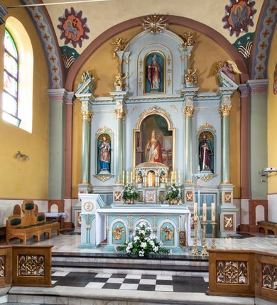 Zdjęcie nr 13: Nastawa ołtarza głównego przyjęła formę przyściennej, trójosiowej, jednokondygnacyjnej struktury architektonicznej ze wieńczeniem. Drewniany stipes ołtarzowy uzyskał kształt leżącego prostopadłościanu podtrzymującego mensę z portatylem, ustawionego na jednostopniowym podeście. Front stipesu udekorowano trzema płycinami obwiedzionymi profilowanymi listwami z rzeźbionymi ornamentami w polach. Na mensie ustawiono drewniane tabernakulum na rzucie trapezu. W dolnej części artykułują je czworoboczne płyciny z ornamentem geometrycznym, rozdzielone spływami wolutowymi podtrzymującymi profilowany gzyms, a w górnej części rozbudowany tron wystawienia. Prostopadłościenne, pancerne tabernakulum usytuowano na osi zdobiąc jego czworoboczne drzwiczki płyciną z trybowanym przedstawieniem kielicha w otoczeniu winnej latorośli i kłosów zbóż. Tron wystawienia przyjął formę trójarkadowej kapliczki zwieńczonej gzymsem koronującym i spłaszczonym dachem pokrytym złoconą rybią łuską. Arkady wsparto na korynckich, kanelowanych kolumienkach. W przyłuczach arkady środkowej, wypełnionej niszą na monstrancję, ukazano uskrzydlone dwie główki anielskie, a w przyłuczach arkad skrajnych motywy ornamentalne. Pod arkadami w osiach skrajnych umieszczono w niszach klęczących na jedno kolana na postumentach adorujących aniołów. Wsparty na arkadach gzyms koronujący ozdobiono profilowaniem i rytmicznie powtarzającymi się konsolkami. Za stipesem ustawiono strukturę ołtarzowa wspartą na wysokim cokole, artykułowaną czterema wysuniętym do przodu dwustopniowymi postumentami dekorowanymi profilowanym gzymsowaniem oraz prostokątnymi płycinami ujętymi w profilowane obramienia. W górnej strefie w płycinach od frontu wyobrażono uskrzydlone głowy symboli czterech Ewangelistów. Idę od lewej jest to głowa woła, anioła, orła i lwa. Natomiast w dolne strefie w płycinach znalazły się od lewej: skrzyżowane bicze z trzciną, na tle których wyobrażono chustę Weroniki, świecznik siedmioramienny z miedzianym wężem na palu na tle pektorału żydowskiego arcykapłana, tablice Dekalogu na tle skrzyżowanych trąb oraz krzyż skrzyżowany z kolumną biczowania wraz z lancą i gąbką na kiju. Pomiędzy postumentami w osiach skrajnych partii cokołowej usytuowano dwoje drzwi prowadzących do zakrystii, zdobionych czworobocznymi płycinami. Na postumentach ustawiono cztery kanelowane kolumny o kapitelach kompozytowych z impostami podtrzymujących belkowanie wyłamane na ich osi. Pomiędzy kolumnami artykułującymi kondygnację w osi środkowej umieszczono pole obrazowe, a w osiach skrajnych półkoliste nisze z ustawionymi w nich na postumentach figurami ŚŚ. Apostołów Piotra i Pawła. Zarówno nad polem obrazowym, jak i nad niszami oraz pod nimi widoczny jest ornament roślinny. Pole obrazowe na osi retabulum przyjęło kształt stojącego prostokąta z nadwieszonym półkolistym łukiem odcinkowym. Ujmuje je profilowana rama. W polu umieszczono obraz Wniebowzięcia Najświętszej Marii Panny, a na dwóch zasuwach przedstawienie św. Urbana oraz św. Barbary. Na belkowaniu w osiach skrajnych nad kolumnami zewnętrznymi usytuowano dwa przerwane przyczółki z leżącymi na nich parą aniołów trzymających krzyż i kotwicę. Natomiast na osi kolumn wewnętrznych na postumentach siedzą dwa złocone putta. Zwieńczenie ołtarza przyjęło kształt leżącego prostokąta flankowanego przez pilaster i kolumienkę podtrzymujących odcinki wyłamanego belkowania, na którym wsparto półkolisty, profilowany łuk odcinkowy wieńczący całość. Ponad tymi podporami umieszczono złocone, siedzące putta, a na osi łuku wieńczącego w promieniach z obłoczkami gołębicę Ducha Świętego. W polu zwieńczenia usytuowano półkolistą niszę ze stojącą figurą Boga Ojca. Ujmuje ją półkoliste obramienie zdobione stylizowaną ornamentyką roślinno-geometryczną. Strukturę architektoniczną retabulum polichromowano w tonacji jasnopastelowej zieleni, ornamenty, profile architektoniczne zostały wyzłocone, natomiast figury apostołów, Boga Ojca oraz aniołów przy tabernakulum i w zwieńczeniu w osiach skrajnych zostały naturalistycznie polichromowane. 