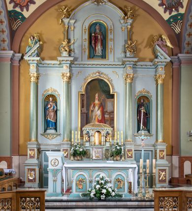 Zdjęcie nr 2: Nastawa ołtarza głównego przyjęła formę przyściennej, trójosiowej, jednokondygnacyjnej struktury architektonicznej ze wieńczeniem. Drewniany stipes ołtarzowy uzyskał kształt leżącego prostopadłościanu podtrzymującego mensę z portatylem, ustawionego na jednostopniowym podeście. Front stipesu udekorowano trzema płycinami obwiedzionymi profilowanymi listwami z rzeźbionymi ornamentami w polach. Na mensie ustawiono drewniane tabernakulum na rzucie trapezu. W dolnej części artykułują je czworoboczne płyciny z ornamentem geometrycznym, rozdzielone spływami wolutowymi podtrzymującymi profilowany gzyms, a w górnej części rozbudowany tron wystawienia. Prostopadłościenne, pancerne tabernakulum usytuowano na osi zdobiąc jego czworoboczne drzwiczki płyciną z trybowanym przedstawieniem kielicha w otoczeniu winnej latorośli i kłosów zbóż. Tron wystawienia przyjął formę trójarkadowej kapliczki zwieńczonej gzymsem koronującym i spłaszczonym dachem pokrytym złoconą rybią łuską. Arkady wsparto na korynckich, kanelowanych kolumienkach. W przyłuczach arkady środkowej, wypełnionej niszą na monstrancję, ukazano uskrzydlone dwie główki anielskie, a w przyłuczach arkad skrajnych motywy ornamentalne. Pod arkadami w osiach skrajnych umieszczono w niszach klęczących na jedno kolana na postumentach adorujących aniołów. Wsparty na arkadach gzyms koronujący ozdobiono profilowaniem i rytmicznie powtarzającymi się konsolkami. Za stipesem ustawiono strukturę ołtarzowa wspartą na wysokim cokole, artykułowaną czterema wysuniętym do przodu dwustopniowymi postumentami dekorowanymi profilowanym gzymsowaniem oraz prostokątnymi płycinami ujętymi w profilowane obramienia. W górnej strefie w płycinach od frontu wyobrażono uskrzydlone głowy symboli czterech Ewangelistów. Idę od lewej jest to głowa woła, anioła, orła i lwa. Natomiast w dolne strefie w płycinach znalazły się od lewej: skrzyżowane bicze z trzciną, na tle których wyobrażono chustę Weroniki, świecznik siedmioramienny z miedzianym wężem na palu na tle pektorału żydowskiego arcykapłana, tablice Dekalogu na tle skrzyżowanych trąb oraz krzyż skrzyżowany z kolumną biczowania wraz z lancą i gąbką na kiju. Pomiędzy postumentami w osiach skrajnych partii cokołowej usytuowano dwoje drzwi prowadzących do zakrystii, zdobionych czworobocznymi płycinami. Na postumentach ustawiono cztery kanelowane kolumny o kapitelach kompozytowych z impostami podtrzymujących belkowanie wyłamane na ich osi. Pomiędzy kolumnami artykułującymi kondygnację w osi środkowej umieszczono pole obrazowe, a w osiach skrajnych półkoliste nisze z ustawionymi w nich na postumentach figurami ŚŚ. Apostołów Piotra i Pawła. Zarówno nad polem obrazowym, jak i nad niszami oraz pod nimi widoczny jest ornament roślinny. Pole obrazowe na osi retabulum przyjęło kształt stojącego prostokąta z nadwieszonym półkolistym łukiem odcinkowym. Ujmuje je profilowana rama. W polu umieszczono obraz Wniebowzięcia Najświętszej Marii Panny, a na dwóch zasuwach przedstawienie św. Urbana oraz św. Barbary. Na belkowaniu w osiach skrajnych nad kolumnami zewnętrznymi usytuowano dwa przerwane przyczółki z leżącymi na nich parą aniołów trzymających krzyż i kotwicę. Natomiast na osi kolumn wewnętrznych na postumentach siedzą dwa złocone putta. Zwieńczenie ołtarza przyjęło kształt leżącego prostokąta flankowanego przez pilaster i kolumienkę podtrzymujących odcinki wyłamanego belkowania, na którym wsparto półkolisty, profilowany łuk odcinkowy wieńczący całość. Ponad tymi podporami umieszczono złocone, siedzące putta, a na osi łuku wieńczącego w promieniach z obłoczkami gołębicę Ducha Świętego. W polu zwieńczenia usytuowano półkolistą niszę ze stojącą figurą Boga Ojca. Ujmuje ją półkoliste obramienie zdobione stylizowaną ornamentyką roślinno-geometryczną. Strukturę architektoniczną retabulum polichromowano w tonacji jasnopastelowej zieleni, ornamenty, profile architektoniczne zostały wyzłocone, natomiast figury apostołów, Boga Ojca oraz aniołów przy tabernakulum i w zwieńczeniu w osiach skrajnych zostały naturalistycznie polichromowane. 