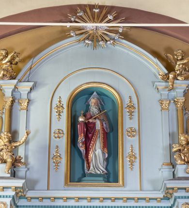 Zdjęcie nr 14: Nastawa ołtarza głównego przyjęła formę przyściennej, trójosiowej, jednokondygnacyjnej struktury architektonicznej ze wieńczeniem. Drewniany stipes ołtarzowy uzyskał kształt leżącego prostopadłościanu podtrzymującego mensę z portatylem, ustawionego na jednostopniowym podeście. Front stipesu udekorowano trzema płycinami obwiedzionymi profilowanymi listwami z rzeźbionymi ornamentami w polach. Na mensie ustawiono drewniane tabernakulum na rzucie trapezu. W dolnej części artykułują je czworoboczne płyciny z ornamentem geometrycznym, rozdzielone spływami wolutowymi podtrzymującymi profilowany gzyms, a w górnej części rozbudowany tron wystawienia. Prostopadłościenne, pancerne tabernakulum usytuowano na osi zdobiąc jego czworoboczne drzwiczki płyciną z trybowanym przedstawieniem kielicha w otoczeniu winnej latorośli i kłosów zbóż. Tron wystawienia przyjął formę trójarkadowej kapliczki zwieńczonej gzymsem koronującym i spłaszczonym dachem pokrytym złoconą rybią łuską. Arkady wsparto na korynckich, kanelowanych kolumienkach. W przyłuczach arkady środkowej, wypełnionej niszą na monstrancję, ukazano uskrzydlone dwie główki anielskie, a w przyłuczach arkad skrajnych motywy ornamentalne. Pod arkadami w osiach skrajnych umieszczono w niszach klęczących na jedno kolana na postumentach adorujących aniołów. Wsparty na arkadach gzyms koronujący ozdobiono profilowaniem i rytmicznie powtarzającymi się konsolkami. Za stipesem ustawiono strukturę ołtarzowa wspartą na wysokim cokole, artykułowaną czterema wysuniętym do przodu dwustopniowymi postumentami dekorowanymi profilowanym gzymsowaniem oraz prostokątnymi płycinami ujętymi w profilowane obramienia. W górnej strefie w płycinach od frontu wyobrażono uskrzydlone głowy symboli czterech Ewangelistów. Idę od lewej jest to głowa woła, anioła, orła i lwa. Natomiast w dolne strefie w płycinach znalazły się od lewej: skrzyżowane bicze z trzciną, na tle których wyobrażono chustę Weroniki, świecznik siedmioramienny z miedzianym wężem na palu na tle pektorału żydowskiego arcykapłana, tablice Dekalogu na tle skrzyżowanych trąb oraz krzyż skrzyżowany z kolumną biczowania wraz z lancą i gąbką na kiju. Pomiędzy postumentami w osiach skrajnych partii cokołowej usytuowano dwoje drzwi prowadzących do zakrystii, zdobionych czworobocznymi płycinami. Na postumentach ustawiono cztery kanelowane kolumny o kapitelach kompozytowych z impostami podtrzymujących belkowanie wyłamane na ich osi. Pomiędzy kolumnami artykułującymi kondygnację w osi środkowej umieszczono pole obrazowe, a w osiach skrajnych półkoliste nisze z ustawionymi w nich na postumentach figurami ŚŚ. Apostołów Piotra i Pawła. Zarówno nad polem obrazowym, jak i nad niszami oraz pod nimi widoczny jest ornament roślinny. Pole obrazowe na osi retabulum przyjęło kształt stojącego prostokąta z nadwieszonym półkolistym łukiem odcinkowym. Ujmuje je profilowana rama. W polu umieszczono obraz Wniebowzięcia Najświętszej Marii Panny, a na dwóch zasuwach przedstawienie św. Urbana oraz św. Barbary. Na belkowaniu w osiach skrajnych nad kolumnami zewnętrznymi usytuowano dwa przerwane przyczółki z leżącymi na nich parą aniołów trzymających krzyż i kotwicę. Natomiast na osi kolumn wewnętrznych na postumentach siedzą dwa złocone putta. Zwieńczenie ołtarza przyjęło kształt leżącego prostokąta flankowanego przez pilaster i kolumienkę podtrzymujących odcinki wyłamanego belkowania, na którym wsparto półkolisty, profilowany łuk odcinkowy wieńczący całość. Ponad tymi podporami umieszczono złocone, siedzące putta, a na osi łuku wieńczącego w promieniach z obłoczkami gołębicę Ducha Świętego. W polu zwieńczenia usytuowano półkolistą niszę ze stojącą figurą Boga Ojca. Ujmuje ją półkoliste obramienie zdobione stylizowaną ornamentyką roślinno-geometryczną. Strukturę architektoniczną retabulum polichromowano w tonacji jasnopastelowej zieleni, ornamenty, profile architektoniczne zostały wyzłocone, natomiast figury apostołów, Boga Ojca oraz aniołów przy tabernakulum i w zwieńczeniu w osiach skrajnych zostały naturalistycznie polichromowane. 