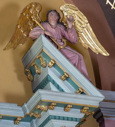 Zdjęcie nr 20: Nastawa ołtarza głównego przyjęła formę przyściennej, trójosiowej, jednokondygnacyjnej struktury architektonicznej ze wieńczeniem. Drewniany stipes ołtarzowy uzyskał kształt leżącego prostopadłościanu podtrzymującego mensę z portatylem, ustawionego na jednostopniowym podeście. Front stipesu udekorowano trzema płycinami obwiedzionymi profilowanymi listwami z rzeźbionymi ornamentami w polach. Na mensie ustawiono drewniane tabernakulum na rzucie trapezu. W dolnej części artykułują je czworoboczne płyciny z ornamentem geometrycznym, rozdzielone spływami wolutowymi podtrzymującymi profilowany gzyms, a w górnej części rozbudowany tron wystawienia. Prostopadłościenne, pancerne tabernakulum usytuowano na osi zdobiąc jego czworoboczne drzwiczki płyciną z trybowanym przedstawieniem kielicha w otoczeniu winnej latorośli i kłosów zbóż. Tron wystawienia przyjął formę trójarkadowej kapliczki zwieńczonej gzymsem koronującym i spłaszczonym dachem pokrytym złoconą rybią łuską. Arkady wsparto na korynckich, kanelowanych kolumienkach. W przyłuczach arkady środkowej, wypełnionej niszą na monstrancję, ukazano uskrzydlone dwie główki anielskie, a w przyłuczach arkad skrajnych motywy ornamentalne. Pod arkadami w osiach skrajnych umieszczono w niszach klęczących na jedno kolana na postumentach adorujących aniołów. Wsparty na arkadach gzyms koronujący ozdobiono profilowaniem i rytmicznie powtarzającymi się konsolkami. Za stipesem ustawiono strukturę ołtarzowa wspartą na wysokim cokole, artykułowaną czterema wysuniętym do przodu dwustopniowymi postumentami dekorowanymi profilowanym gzymsowaniem oraz prostokątnymi płycinami ujętymi w profilowane obramienia. W górnej strefie w płycinach od frontu wyobrażono uskrzydlone głowy symboli czterech Ewangelistów. Idę od lewej jest to głowa woła, anioła, orła i lwa. Natomiast w dolne strefie w płycinach znalazły się od lewej: skrzyżowane bicze z trzciną, na tle których wyobrażono chustę Weroniki, świecznik siedmioramienny z miedzianym wężem na palu na tle pektorału żydowskiego arcykapłana, tablice Dekalogu na tle skrzyżowanych trąb oraz krzyż skrzyżowany z kolumną biczowania wraz z lancą i gąbką na kiju. Pomiędzy postumentami w osiach skrajnych partii cokołowej usytuowano dwoje drzwi prowadzących do zakrystii, zdobionych czworobocznymi płycinami. Na postumentach ustawiono cztery kanelowane kolumny o kapitelach kompozytowych z impostami podtrzymujących belkowanie wyłamane na ich osi. Pomiędzy kolumnami artykułującymi kondygnację w osi środkowej umieszczono pole obrazowe, a w osiach skrajnych półkoliste nisze z ustawionymi w nich na postumentach figurami ŚŚ. Apostołów Piotra i Pawła. Zarówno nad polem obrazowym, jak i nad niszami oraz pod nimi widoczny jest ornament roślinny. Pole obrazowe na osi retabulum przyjęło kształt stojącego prostokąta z nadwieszonym półkolistym łukiem odcinkowym. Ujmuje je profilowana rama. W polu umieszczono obraz Wniebowzięcia Najświętszej Marii Panny, a na dwóch zasuwach przedstawienie św. Urbana oraz św. Barbary. Na belkowaniu w osiach skrajnych nad kolumnami zewnętrznymi usytuowano dwa przerwane przyczółki z leżącymi na nich parą aniołów trzymających krzyż i kotwicę. Natomiast na osi kolumn wewnętrznych na postumentach siedzą dwa złocone putta. Zwieńczenie ołtarza przyjęło kształt leżącego prostokąta flankowanego przez pilaster i kolumienkę podtrzymujących odcinki wyłamanego belkowania, na którym wsparto półkolisty, profilowany łuk odcinkowy wieńczący całość. Ponad tymi podporami umieszczono złocone, siedzące putta, a na osi łuku wieńczącego w promieniach z obłoczkami gołębicę Ducha Świętego. W polu zwieńczenia usytuowano półkolistą niszę ze stojącą figurą Boga Ojca. Ujmuje ją półkoliste obramienie zdobione stylizowaną ornamentyką roślinno-geometryczną. Strukturę architektoniczną retabulum polichromowano w tonacji jasnopastelowej zieleni, ornamenty, profile architektoniczne zostały wyzłocone, natomiast figury apostołów, Boga Ojca oraz aniołów przy tabernakulum i w zwieńczeniu w osiach skrajnych zostały naturalistycznie polichromowane. 