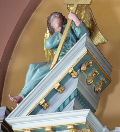 Zdjęcie nr 19: Nastawa ołtarza głównego przyjęła formę przyściennej, trójosiowej, jednokondygnacyjnej struktury architektonicznej ze wieńczeniem. Drewniany stipes ołtarzowy uzyskał kształt leżącego prostopadłościanu podtrzymującego mensę z portatylem, ustawionego na jednostopniowym podeście. Front stipesu udekorowano trzema płycinami obwiedzionymi profilowanymi listwami z rzeźbionymi ornamentami w polach. Na mensie ustawiono drewniane tabernakulum na rzucie trapezu. W dolnej części artykułują je czworoboczne płyciny z ornamentem geometrycznym, rozdzielone spływami wolutowymi podtrzymującymi profilowany gzyms, a w górnej części rozbudowany tron wystawienia. Prostopadłościenne, pancerne tabernakulum usytuowano na osi zdobiąc jego czworoboczne drzwiczki płyciną z trybowanym przedstawieniem kielicha w otoczeniu winnej latorośli i kłosów zbóż. Tron wystawienia przyjął formę trójarkadowej kapliczki zwieńczonej gzymsem koronującym i spłaszczonym dachem pokrytym złoconą rybią łuską. Arkady wsparto na korynckich, kanelowanych kolumienkach. W przyłuczach arkady środkowej, wypełnionej niszą na monstrancję, ukazano uskrzydlone dwie główki anielskie, a w przyłuczach arkad skrajnych motywy ornamentalne. Pod arkadami w osiach skrajnych umieszczono w niszach klęczących na jedno kolana na postumentach adorujących aniołów. Wsparty na arkadach gzyms koronujący ozdobiono profilowaniem i rytmicznie powtarzającymi się konsolkami. Za stipesem ustawiono strukturę ołtarzowa wspartą na wysokim cokole, artykułowaną czterema wysuniętym do przodu dwustopniowymi postumentami dekorowanymi profilowanym gzymsowaniem oraz prostokątnymi płycinami ujętymi w profilowane obramienia. W górnej strefie w płycinach od frontu wyobrażono uskrzydlone głowy symboli czterech Ewangelistów. Idę od lewej jest to głowa woła, anioła, orła i lwa. Natomiast w dolne strefie w płycinach znalazły się od lewej: skrzyżowane bicze z trzciną, na tle których wyobrażono chustę Weroniki, świecznik siedmioramienny z miedzianym wężem na palu na tle pektorału żydowskiego arcykapłana, tablice Dekalogu na tle skrzyżowanych trąb oraz krzyż skrzyżowany z kolumną biczowania wraz z lancą i gąbką na kiju. Pomiędzy postumentami w osiach skrajnych partii cokołowej usytuowano dwoje drzwi prowadzących do zakrystii, zdobionych czworobocznymi płycinami. Na postumentach ustawiono cztery kanelowane kolumny o kapitelach kompozytowych z impostami podtrzymujących belkowanie wyłamane na ich osi. Pomiędzy kolumnami artykułującymi kondygnację w osi środkowej umieszczono pole obrazowe, a w osiach skrajnych półkoliste nisze z ustawionymi w nich na postumentach figurami ŚŚ. Apostołów Piotra i Pawła. Zarówno nad polem obrazowym, jak i nad niszami oraz pod nimi widoczny jest ornament roślinny. Pole obrazowe na osi retabulum przyjęło kształt stojącego prostokąta z nadwieszonym półkolistym łukiem odcinkowym. Ujmuje je profilowana rama. W polu umieszczono obraz Wniebowzięcia Najświętszej Marii Panny, a na dwóch zasuwach przedstawienie św. Urbana oraz św. Barbary. Na belkowaniu w osiach skrajnych nad kolumnami zewnętrznymi usytuowano dwa przerwane przyczółki z leżącymi na nich parą aniołów trzymających krzyż i kotwicę. Natomiast na osi kolumn wewnętrznych na postumentach siedzą dwa złocone putta. Zwieńczenie ołtarza przyjęło kształt leżącego prostokąta flankowanego przez pilaster i kolumienkę podtrzymujących odcinki wyłamanego belkowania, na którym wsparto półkolisty, profilowany łuk odcinkowy wieńczący całość. Ponad tymi podporami umieszczono złocone, siedzące putta, a na osi łuku wieńczącego w promieniach z obłoczkami gołębicę Ducha Świętego. W polu zwieńczenia usytuowano półkolistą niszę ze stojącą figurą Boga Ojca. Ujmuje ją półkoliste obramienie zdobione stylizowaną ornamentyką roślinno-geometryczną. Strukturę architektoniczną retabulum polichromowano w tonacji jasnopastelowej zieleni, ornamenty, profile architektoniczne zostały wyzłocone, natomiast figury apostołów, Boga Ojca oraz aniołów przy tabernakulum i w zwieńczeniu w osiach skrajnych zostały naturalistycznie polichromowane. 