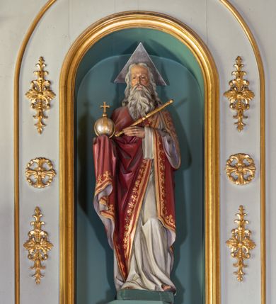 Zdjęcie nr 15: Nastawa ołtarza głównego przyjęła formę przyściennej, trójosiowej, jednokondygnacyjnej struktury architektonicznej ze wieńczeniem. Drewniany stipes ołtarzowy uzyskał kształt leżącego prostopadłościanu podtrzymującego mensę z portatylem, ustawionego na jednostopniowym podeście. Front stipesu udekorowano trzema płycinami obwiedzionymi profilowanymi listwami z rzeźbionymi ornamentami w polach. Na mensie ustawiono drewniane tabernakulum na rzucie trapezu. W dolnej części artykułują je czworoboczne płyciny z ornamentem geometrycznym, rozdzielone spływami wolutowymi podtrzymującymi profilowany gzyms, a w górnej części rozbudowany tron wystawienia. Prostopadłościenne, pancerne tabernakulum usytuowano na osi zdobiąc jego czworoboczne drzwiczki płyciną z trybowanym przedstawieniem kielicha w otoczeniu winnej latorośli i kłosów zbóż. Tron wystawienia przyjął formę trójarkadowej kapliczki zwieńczonej gzymsem koronującym i spłaszczonym dachem pokrytym złoconą rybią łuską. Arkady wsparto na korynckich, kanelowanych kolumienkach. W przyłuczach arkady środkowej, wypełnionej niszą na monstrancję, ukazano uskrzydlone dwie główki anielskie, a w przyłuczach arkad skrajnych motywy ornamentalne. Pod arkadami w osiach skrajnych umieszczono w niszach klęczących na jedno kolana na postumentach adorujących aniołów. Wsparty na arkadach gzyms koronujący ozdobiono profilowaniem i rytmicznie powtarzającymi się konsolkami. Za stipesem ustawiono strukturę ołtarzowa wspartą na wysokim cokole, artykułowaną czterema wysuniętym do przodu dwustopniowymi postumentami dekorowanymi profilowanym gzymsowaniem oraz prostokątnymi płycinami ujętymi w profilowane obramienia. W górnej strefie w płycinach od frontu wyobrażono uskrzydlone głowy symboli czterech Ewangelistów. Idę od lewej jest to głowa woła, anioła, orła i lwa. Natomiast w dolne strefie w płycinach znalazły się od lewej: skrzyżowane bicze z trzciną, na tle których wyobrażono chustę Weroniki, świecznik siedmioramienny z miedzianym wężem na palu na tle pektorału żydowskiego arcykapłana, tablice Dekalogu na tle skrzyżowanych trąb oraz krzyż skrzyżowany z kolumną biczowania wraz z lancą i gąbką na kiju. Pomiędzy postumentami w osiach skrajnych partii cokołowej usytuowano dwoje drzwi prowadzących do zakrystii, zdobionych czworobocznymi płycinami. Na postumentach ustawiono cztery kanelowane kolumny o kapitelach kompozytowych z impostami podtrzymujących belkowanie wyłamane na ich osi. Pomiędzy kolumnami artykułującymi kondygnację w osi środkowej umieszczono pole obrazowe, a w osiach skrajnych półkoliste nisze z ustawionymi w nich na postumentach figurami ŚŚ. Apostołów Piotra i Pawła. Zarówno nad polem obrazowym, jak i nad niszami oraz pod nimi widoczny jest ornament roślinny. Pole obrazowe na osi retabulum przyjęło kształt stojącego prostokąta z nadwieszonym półkolistym łukiem odcinkowym. Ujmuje je profilowana rama. W polu umieszczono obraz Wniebowzięcia Najświętszej Marii Panny, a na dwóch zasuwach przedstawienie św. Urbana oraz św. Barbary. Na belkowaniu w osiach skrajnych nad kolumnami zewnętrznymi usytuowano dwa przerwane przyczółki z leżącymi na nich parą aniołów trzymających krzyż i kotwicę. Natomiast na osi kolumn wewnętrznych na postumentach siedzą dwa złocone putta. Zwieńczenie ołtarza przyjęło kształt leżącego prostokąta flankowanego przez pilaster i kolumienkę podtrzymujących odcinki wyłamanego belkowania, na którym wsparto półkolisty, profilowany łuk odcinkowy wieńczący całość. Ponad tymi podporami umieszczono złocone, siedzące putta, a na osi łuku wieńczącego w promieniach z obłoczkami gołębicę Ducha Świętego. W polu zwieńczenia usytuowano półkolistą niszę ze stojącą figurą Boga Ojca. Ujmuje ją półkoliste obramienie zdobione stylizowaną ornamentyką roślinno-geometryczną. Strukturę architektoniczną retabulum polichromowano w tonacji jasnopastelowej zieleni, ornamenty, profile architektoniczne zostały wyzłocone, natomiast figury apostołów, Boga Ojca oraz aniołów przy tabernakulum i w zwieńczeniu w osiach skrajnych zostały naturalistycznie polichromowane. 