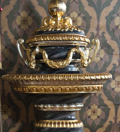 Zdjęcie nr 1: Chrzcielnica w typie kielichowym osadzona na czworobocznej, profilowanej, zwężającej się ku górze stopie. Niewysoki trzon jest w formie kanelurowanego trzonu czworobocznego filara. Powyżej wąski, puklowany nodus, nad nim jest wklęska ujęta od góry pierścieniem z motywem astragalu. W górnej partii przybiera formę rozszerzającej się, profilowanej i zrobionej motywem rozetek profitki o wywiniętej na zewnątrz krawędzi zdobionej motywem lancetowatych liści. Misa chrzcielnicy przybiera formę owalnej wazy o lekko spłaszczonym brzuścu ujętym parą wolutowych imadeł. U ujętego wałkiem wylewu podwieszony w dwóch miejscach rodzaj festonu z liści laurowych. Pokrywa zwieńczona kompozycją liści i pary wolut wspierających kulę z krzyżem. Struktura chrzcielnicy marmoryzowana w odcieniach szarości, ornamentyka złocona i srebrzona.  