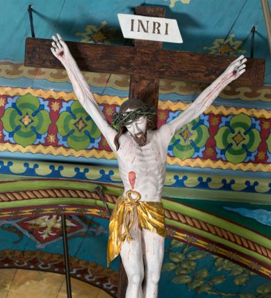 Zdjęcie nr 1: Figura Jezusa Ukrzyżowanego na belce tęczowej przedstawia typ Cristo morte. Utrzymane w sinobladej kolorystyce ciało Jezusa, przybite trzema gwoździami, charakteryzuje wydatna, stosunkowo sumarycznie opracowana muskulatura z wyraźnie zaakcentowanymi układami ścięgien i żył. Opadającą na prawy bark głowę cechuje pociągła twarz o sumarycznie opracowanych rysach, z długim, szerokim nosem oraz krótkim, ciemnobrązowym zarostem. Falowane włosy opadają na plecy, z jednym puklem na prawym ramieniu. Głowę wieńczy szeroka korona cierniowa. Ukazane w asymetrycznym układzie ramiona są w głębokim, V-kształtnym zwisie. Wydatną klatkę piersiową akcentują wyraziste linie żeber. Nogi są lekko zgięte w kolanach, a stopy szerokie. Biodra osłania krótkie, złocone perizonium przewiązane sznurem na prawym boku. W prawym boku Jezusa widoczna jest wyrazista, krwawiąca rana, sylwetkę pokrywają drobne stróżki krwi. W górnej partii krzyża znajduje się romboidalny titulus z napisem „INRI”.