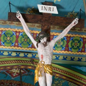 Zdjęcie nr 1: Figura Jezusa Ukrzyżowanego na belce tęczowej przedstawia typ Cristo morte. Utrzymane w sinobladej kolorystyce ciało Jezusa, przybite trzema gwoździami, charakteryzuje wydatna, stosunkowo sumarycznie opracowana muskulatura z wyraźnie zaakcentowanymi układami ścięgien i żył. Opadającą na prawy bark głowę cechuje pociągła twarz o sumarycznie opracowanych rysach, z długim, szerokim nosem oraz krótkim, ciemnobrązowym zarostem. Falowane włosy opadają na plecy, z jednym puklem na prawym ramieniu. Głowę wieńczy szeroka korona cierniowa. Ukazane w asymetrycznym układzie ramiona są w głębokim, V-kształtnym zwisie. Wydatną klatkę piersiową akcentują wyraziste linie żeber. Nogi są lekko zgięte w kolanach, a stopy szerokie. Biodra osłania krótkie, złocone perizonium przewiązane sznurem na prawym boku. W prawym boku Jezusa widoczna jest wyrazista, krwawiąca rana, sylwetkę pokrywają drobne stróżki krwi. W górnej partii krzyża znajduje się romboidalny titulus z napisem „INRI”.