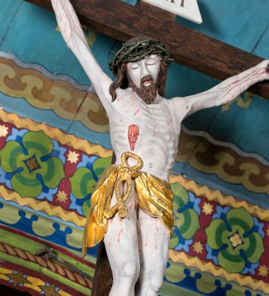 Zdjęcie nr 3: Figura Jezusa Ukrzyżowanego na belce tęczowej przedstawia typ Cristo morte. Utrzymane w sinobladej kolorystyce ciało Jezusa, przybite trzema gwoździami, charakteryzuje wydatna, stosunkowo sumarycznie opracowana muskulatura z wyraźnie zaakcentowanymi układami ścięgien i żył. Opadającą na prawy bark głowę cechuje pociągła twarz o sumarycznie opracowanych rysach, z długim, szerokim nosem oraz krótkim, ciemnobrązowym zarostem. Falowane włosy opadają na plecy, z jednym puklem na prawym ramieniu. Głowę wieńczy szeroka korona cierniowa. Ukazane w asymetrycznym układzie ramiona są w głębokim, V-kształtnym zwisie. Wydatną klatkę piersiową akcentują wyraziste linie żeber. Nogi są lekko zgięte w kolanach, a stopy szerokie. Biodra osłania krótkie, złocone perizonium przewiązane sznurem na prawym boku. W prawym boku Jezusa widoczna jest wyrazista, krwawiąca rana, sylwetkę pokrywają drobne stróżki krwi. W górnej partii krzyża znajduje się romboidalny titulus z napisem „INRI”.