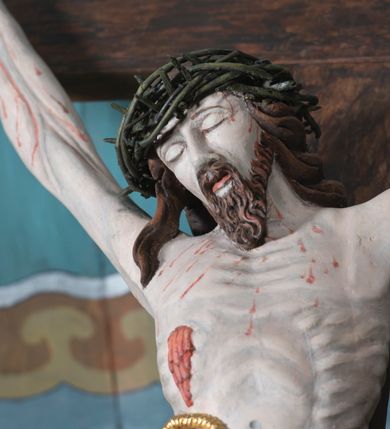 Zdjęcie nr 2: Figura Jezusa Ukrzyżowanego na belce tęczowej przedstawia typ Cristo morte. Utrzymane w sinobladej kolorystyce ciało Jezusa, przybite trzema gwoździami, charakteryzuje wydatna, stosunkowo sumarycznie opracowana muskulatura z wyraźnie zaakcentowanymi układami ścięgien i żył. Opadającą na prawy bark głowę cechuje pociągła twarz o sumarycznie opracowanych rysach, z długim, szerokim nosem oraz krótkim, ciemnobrązowym zarostem. Falowane włosy opadają na plecy, z jednym puklem na prawym ramieniu. Głowę wieńczy szeroka korona cierniowa. Ukazane w asymetrycznym układzie ramiona są w głębokim, V-kształtnym zwisie. Wydatną klatkę piersiową akcentują wyraziste linie żeber. Nogi są lekko zgięte w kolanach, a stopy szerokie. Biodra osłania krótkie, złocone perizonium przewiązane sznurem na prawym boku. W prawym boku Jezusa widoczna jest wyrazista, krwawiąca rana, sylwetkę pokrywają drobne stróżki krwi. W górnej partii krzyża znajduje się romboidalny titulus z napisem „INRI”.