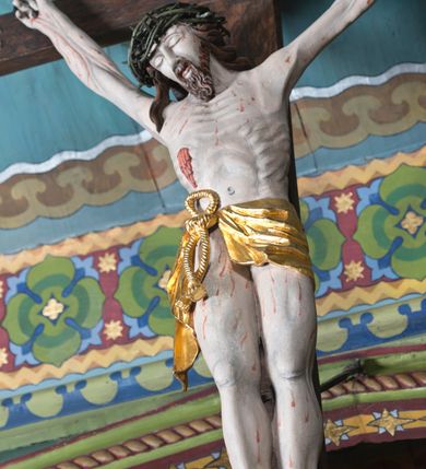 Zdjęcie nr 4: Figura Jezusa Ukrzyżowanego na belce tęczowej przedstawia typ Cristo morte. Utrzymane w sinobladej kolorystyce ciało Jezusa, przybite trzema gwoździami, charakteryzuje wydatna, stosunkowo sumarycznie opracowana muskulatura z wyraźnie zaakcentowanymi układami ścięgien i żył. Opadającą na prawy bark głowę cechuje pociągła twarz o sumarycznie opracowanych rysach, z długim, szerokim nosem oraz krótkim, ciemnobrązowym zarostem. Falowane włosy opadają na plecy, z jednym puklem na prawym ramieniu. Głowę wieńczy szeroka korona cierniowa. Ukazane w asymetrycznym układzie ramiona są w głębokim, V-kształtnym zwisie. Wydatną klatkę piersiową akcentują wyraziste linie żeber. Nogi są lekko zgięte w kolanach, a stopy szerokie. Biodra osłania krótkie, złocone perizonium przewiązane sznurem na prawym boku. W prawym boku Jezusa widoczna jest wyrazista, krwawiąca rana, sylwetkę pokrywają drobne stróżki krwi. W górnej partii krzyża znajduje się romboidalny titulus z napisem „INRI”.