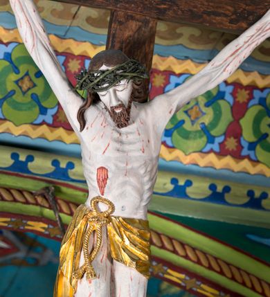 Zdjęcie nr 5: Figura Jezusa Ukrzyżowanego na belce tęczowej przedstawia typ Cristo morte. Utrzymane w sinobladej kolorystyce ciało Jezusa, przybite trzema gwoździami, charakteryzuje wydatna, stosunkowo sumarycznie opracowana muskulatura z wyraźnie zaakcentowanymi układami ścięgien i żył. Opadającą na prawy bark głowę cechuje pociągła twarz o sumarycznie opracowanych rysach, z długim, szerokim nosem oraz krótkim, ciemnobrązowym zarostem. Falowane włosy opadają na plecy, z jednym puklem na prawym ramieniu. Głowę wieńczy szeroka korona cierniowa. Ukazane w asymetrycznym układzie ramiona są w głębokim, V-kształtnym zwisie. Wydatną klatkę piersiową akcentują wyraziste linie żeber. Nogi są lekko zgięte w kolanach, a stopy szerokie. Biodra osłania krótkie, złocone perizonium przewiązane sznurem na prawym boku. W prawym boku Jezusa widoczna jest wyrazista, krwawiąca rana, sylwetkę pokrywają drobne stróżki krwi. W górnej partii krzyża znajduje się romboidalny titulus z napisem „INRI”.