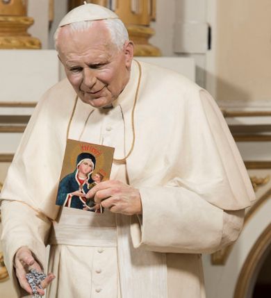 Zdjęcie nr 1: Pełnoplastyczna figura przedstawia św. Jana Pawła II ukazanego w całej postaci, w postawie stojącej i zwróconego na wprost. Papież ukazany jest jako mężczyzna w podeszłym wieku, o przygarbionej sylwetce. Pokrytą głębokimi bruzdami twarz charakteryzują niezwykle realistyczne rysy obrazujące lekki, ciepły uśmiech. Jan Paweł II ubrany jest w papieską sutannę przewiązaną szerokim, jedwabnym pasem z herbem papieskim. Na ramionach ma białą pelerynę, a na głowie piuskę. W lewej, przysuniętej do piersi ręce trzyma niewielki wizerunek Matki Boskiej Kęckiej, w prawej, nieco opuszczonej srebrny różaniec. Figura ustawiona jest na niewysokim, ośmiobocznym postumencie. 