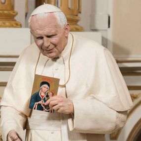 Zdjęcie nr 1: Pełnoplastyczna figura przedstawia św. Jana Pawła II ukazanego w całej postaci, w postawie stojącej i zwróconego na wprost. Papież ukazany jest jako mężczyzna w podeszłym wieku, o przygarbionej sylwetce. Pokrytą głębokimi bruzdami twarz charakteryzują niezwykle realistyczne rysy obrazujące lekki, ciepły uśmiech. Jan Paweł II ubrany jest w papieską sutannę przewiązaną szerokim, jedwabnym pasem z herbem papieskim. Na ramionach ma białą pelerynę, a na głowie piuskę. W lewej, przysuniętej do piersi ręce trzyma niewielki wizerunek Matki Boskiej Kęckiej, w prawej, nieco opuszczonej srebrny różaniec. Figura ustawiona jest na niewysokim, ośmiobocznym postumencie. 