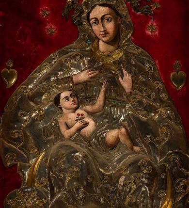 Zdjęcie nr 2: Obraz w kształcie stojącego prostokąta o wklęsło-wypukłej podstawie, zamkniętego nadwieszonym łukiem półkolistym, ujęty profilowaną, złoconą ramą przedstawia tronującą  Madonnę z Dzieciątkiem. Maria zwrócona jest na wprost o nieznacznie przechylonej w lewo sylwetce. Skierowaną w trzech czwartych w prawo, lekko przechyloną głowę charakteryzuje owalna twarz o silnym modelunku światłocieniowym, wyrazistych łukach brwiowych, dużych oczach, długim, prostym nosie i wąskiej linii ust  w odcieniu koralowym. Ciemne włosy rozdzielone są pośrodku głowy przedziałkiem. Partie szat Marii pokrywa metalowa osłona. Matka Boska ma na sobie srebrzoną suknię spiętą kameryzowanym paskiem, pod szyją jest zawiązana złocona chusta, a głowę, ramiona i nogi osłania srebrzony płaszcz zdobiony złoconym motywem wici roślinnej. Głowę Marii wieńczy zamknięta, kameryzowana korona podtrzymywana przez parę unoszących się po bokach aniołów. Prawą rękę składa na piersi, lewą unosi z wyprostowanym palcem wskazującym. Na kolanach Matki Boskiej leży zwrócone w lewo i spoglądające na nią Dzieciątko. Zaokrągloną twarz charakteryzują wyraziste łuki brwi, duże, ciemne oczy, lekko zadarty nos, kształtne usta i wyraźnie zaznaczony podbródek. Nagą sylwetkę Jezusa okrywa w partii bioder pokryte metalową osłoną, srebrzone perizonium. W prawej, złożonej na piersi dłoni trzyma gałązkę z dwoma kwiatkami czerwonej róży, lewą rękę wyciąga do góry, w kierunku dłoni Marii. Pod stopami Matki Boskiej jest złocony sierp półksiężyca, z tarczą słoneczną w formie rozety po lewej stronie kompozycji, poniżej srebrzone obłoki. Głowę Madonny okala krąg 12 gwiazd, po bokach, na wysokości ramion jest para wotów w kształcie serca. Tło wyłożone jest czerwonym aksamitem.   