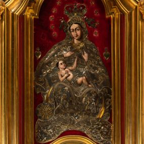 Zdjęcie nr 1: Obraz w kształcie stojącego prostokąta o wklęsło-wypukłej podstawie, zamkniętego nadwieszonym łukiem półkolistym, ujęty profilowaną, złoconą ramą przedstawia tronującą  Madonnę z Dzieciątkiem. Maria zwrócona jest na wprost o nieznacznie przechylonej w lewo sylwetce. Skierowaną w trzech czwartych w prawo, lekko przechyloną głowę charakteryzuje owalna twarz o silnym modelunku światłocieniowym, wyrazistych łukach brwiowych, dużych oczach, długim, prostym nosie i wąskiej linii ust  w odcieniu koralowym. Ciemne włosy rozdzielone są pośrodku głowy przedziałkiem. Partie szat Marii pokrywa metalowa osłona. Matka Boska ma na sobie srebrzoną suknię spiętą kameryzowanym paskiem, pod szyją jest zawiązana złocona chusta, a głowę, ramiona i nogi osłania srebrzony płaszcz zdobiony złoconym motywem wici roślinnej. Głowę Marii wieńczy zamknięta, kameryzowana korona podtrzymywana przez parę unoszących się po bokach aniołów. Prawą rękę składa na piersi, lewą unosi z wyprostowanym palcem wskazującym. Na kolanach Matki Boskiej leży zwrócone w lewo i spoglądające na nią Dzieciątko. Zaokrągloną twarz charakteryzują wyraziste łuki brwi, duże, ciemne oczy, lekko zadarty nos, kształtne usta i wyraźnie zaznaczony podbródek. Nagą sylwetkę Jezusa okrywa w partii bioder pokryte metalową osłoną, srebrzone perizonium. W prawej, złożonej na piersi dłoni trzyma gałązkę z dwoma kwiatkami czerwonej róży, lewą rękę wyciąga do góry, w kierunku dłoni Marii. Pod stopami Matki Boskiej jest złocony sierp półksiężyca, z tarczą słoneczną w formie rozety po lewej stronie kompozycji, poniżej srebrzone obłoki. Głowę Madonny okala krąg 12 gwiazd, po bokach, na wysokości ramion jest para wotów w kształcie serca. Tło wyłożone jest czerwonym aksamitem.   