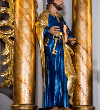 Zdjęcie nr 3: Drążona z tyłu, całopostaciowa figura przedstawia św. Piotra stojącego na niewysokiej podstawie i zwróconego na wprost. Skierowaną w trzech czwartych w lewo głowę charakteryzuje owalna twarz o wyrazistych, sumarycznie opracowanych rysach z długim spiczastym nosem, wystającymi kośćmi policzkowymi oraz krótkim zarostem. Włosy są krótkie z parą zakoli po bokach. Święty ubrany jest w ciemnoniebieską suknię ze złoconymi paskiem i kołnierzem oraz złocony płaszcz narzucony na prawe ramię i podwinięty pod pasek. Spod sukni widoczne bose stopy. W prawej ręce trzyma dwa złocone klucze, lewą podtrzymuje zamkniętą księgę w czerwonej oprawie.     