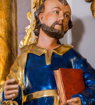 Zdjęcie nr 4: Drążona z tyłu, całopostaciowa figura przedstawia św. Piotra stojącego na niewysokiej podstawie i zwróconego na wprost. Skierowaną w trzech czwartych w lewo głowę charakteryzuje owalna twarz o wyrazistych, sumarycznie opracowanych rysach z długim spiczastym nosem, wystającymi kośćmi policzkowymi oraz krótkim zarostem. Włosy są krótkie z parą zakoli po bokach. Święty ubrany jest w ciemnoniebieską suknię ze złoconymi paskiem i kołnierzem oraz złocony płaszcz narzucony na prawe ramię i podwinięty pod pasek. Spod sukni widoczne bose stopy. W prawej ręce trzyma dwa złocone klucze, lewą podtrzymuje zamkniętą księgę w czerwonej oprawie.     