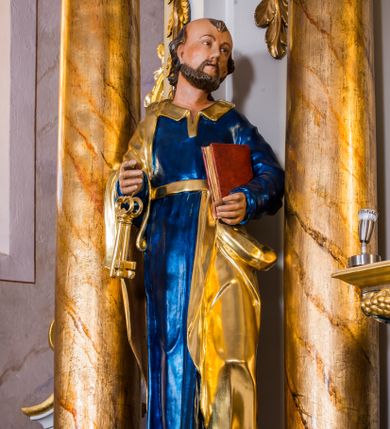 Zdjęcie nr 1: Drążona z tyłu, całopostaciowa figura przedstawia św. Piotra stojącego na niewysokiej podstawie i zwróconego na wprost. Skierowaną w trzech czwartych w lewo głowę charakteryzuje owalna twarz o wyrazistych, sumarycznie opracowanych rysach z długim spiczastym nosem, wystającymi kośćmi policzkowymi oraz krótkim zarostem. Włosy są krótkie z parą zakoli po bokach. Święty ubrany jest w ciemnoniebieską suknię ze złoconymi paskiem i kołnierzem oraz złocony płaszcz narzucony na prawe ramię i podwinięty pod pasek. Spod sukni widoczne bose stopy. W prawej ręce trzyma dwa złocone klucze, lewą podtrzymuje zamkniętą księgę w czerwonej oprawie.     