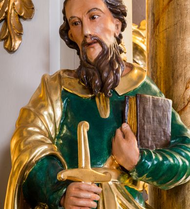 Zdjęcie nr 4: Drążona z tyłu, całopostaciowa figura przedstawia św. Pawła stojącego na niewysokiej podstawie, zwróconego na wprost. Skierowaną w trzech czwartych w prawo głowę charakteryzuje pociągła twarz o wyrazistych, sumarycznie opracowanych rysach z długim spiczastym nosem, wystającymi kośćmi policzkowymi oraz długim zarostem. Lekko skręcone, zebrane do tyłu włosy sięgają ramion. Święty ubrany jest w ciemnozieloną suknię ze złoconymi paskiem i kołnierzem oraz złocony płaszcz narzucony na prawe ramię i podwinięty pod pasek. W prawej ręce trzyma złocono-srebrzony miecz skierowany ostrzem w dół, lewą przyciska do piersi zamkniętą księgę w brązowej oprawie.     