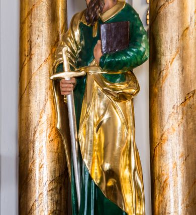 Zdjęcie nr 3: Drążona z tyłu, całopostaciowa figura przedstawia św. Pawła stojącego na niewysokiej podstawie, zwróconego na wprost. Skierowaną w trzech czwartych w prawo głowę charakteryzuje pociągła twarz o wyrazistych, sumarycznie opracowanych rysach z długim spiczastym nosem, wystającymi kośćmi policzkowymi oraz długim zarostem. Lekko skręcone, zebrane do tyłu włosy sięgają ramion. Święty ubrany jest w ciemnozieloną suknię ze złoconymi paskiem i kołnierzem oraz złocony płaszcz narzucony na prawe ramię i podwinięty pod pasek. W prawej ręce trzyma złocono-srebrzony miecz skierowany ostrzem w dół, lewą przyciska do piersi zamkniętą księgę w brązowej oprawie.     