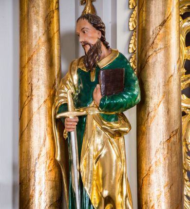 Zdjęcie nr 1: Drążona z tyłu, całopostaciowa figura przedstawia św. Pawła stojącego na niewysokiej podstawie, zwróconego na wprost. Skierowaną w trzech czwartych w prawo głowę charakteryzuje pociągła twarz o wyrazistych, sumarycznie opracowanych rysach z długim spiczastym nosem, wystającymi kośćmi policzkowymi oraz długim zarostem. Lekko skręcone, zebrane do tyłu włosy sięgają ramion. Święty ubrany jest w ciemnozieloną suknię ze złoconymi paskiem i kołnierzem oraz złocony płaszcz narzucony na prawe ramię i podwinięty pod pasek. W prawej ręce trzyma złocono-srebrzony miecz skierowany ostrzem w dół, lewą przyciska do piersi zamkniętą księgę w brązowej oprawie.     