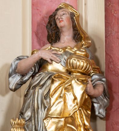 Zdjęcie nr 1: Figura drążona z tyłu, ustawiona na nieregularnej podstawie. Święta zwrócona w trzech czwartych w prawo, w kontrapoście, odchylona łukowato, z głową skierowaną w górę. Prawą dłoń kładzie na piersi, lewą ręką podtrzymuje przy boku płaszcz i puszkę. Twarz ma pociągłą, o podkreślonej brodzie, dużych, głęboko osadzonych, półprzymkniętych oczach, garbatym nosie i małych, pełnych ustach. Włosy długie, gęste, falowane. Ubrana jest w srebrzoną, obfitą i silnie drapowaną suknię o szerokich rękawach, odsłaniającą prawe ramię, przepasaną w talii złoconym gorsetem. Na lewą stronę głowy ma narzucony złocony płaszcz, opadający na lewy bok, silnie drapowany. Polichromia ciała naturalistyczna. 


