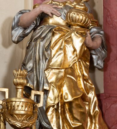 Zdjęcie nr 2: Figura drążona z tyłu, ustawiona na nieregularnej podstawie. Święta zwrócona w trzech czwartych w prawo, w kontrapoście, odchylona łukowato, z głową skierowaną w górę. Prawą dłoń kładzie na piersi, lewą ręką podtrzymuje przy boku płaszcz i puszkę. Twarz ma pociągłą, o podkreślonej brodzie, dużych, głęboko osadzonych, półprzymkniętych oczach, garbatym nosie i małych, pełnych ustach. Włosy długie, gęste, falowane. Ubrana jest w srebrzoną, obfitą i silnie drapowaną suknię o szerokich rękawach, odsłaniającą prawe ramię, przepasaną w talii złoconym gorsetem. Na lewą stronę głowy ma narzucony złocony płaszcz, opadający na lewy bok, silnie drapowany. Polichromia ciała naturalistyczna. 


