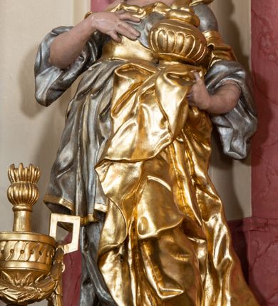 Zdjęcie nr 5: Figura drążona z tyłu, ustawiona na nieregularnej podstawie. Święta zwrócona w trzech czwartych w prawo, w kontrapoście, odchylona łukowato, z głową skierowaną w górę. Prawą dłoń kładzie na piersi, lewą ręką podtrzymuje przy boku płaszcz i puszkę. Twarz ma pociągłą, o podkreślonej brodzie, dużych, głęboko osadzonych, półprzymkniętych oczach, garbatym nosie i małych, pełnych ustach. Włosy długie, gęste, falowane. Ubrana jest w srebrzoną, obfitą i silnie drapowaną suknię o szerokich rękawach, odsłaniającą prawe ramię, przepasaną w talii złoconym gorsetem. Na lewą stronę głowy ma narzucony złocony płaszcz, opadający na lewy bok, silnie drapowany. Polichromia ciała naturalistyczna. 


