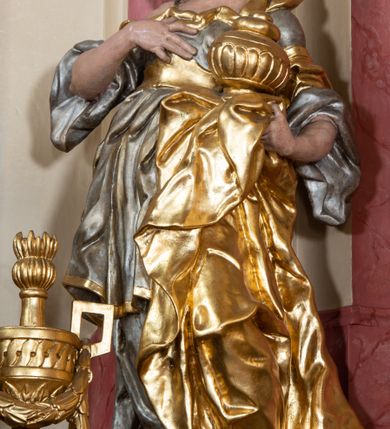 Zdjęcie nr 4: Figura drążona z tyłu, ustawiona na nieregularnej podstawie. Święta zwrócona w trzech czwartych w prawo, w kontrapoście, odchylona łukowato, z głową skierowaną w górę. Prawą dłoń kładzie na piersi, lewą ręką podtrzymuje przy boku płaszcz i puszkę. Twarz ma pociągłą, o podkreślonej brodzie, dużych, głęboko osadzonych, półprzymkniętych oczach, garbatym nosie i małych, pełnych ustach. Włosy długie, gęste, falowane. Ubrana jest w srebrzoną, obfitą i silnie drapowaną suknię o szerokich rękawach, odsłaniającą prawe ramię, przepasaną w talii złoconym gorsetem. Na lewą stronę głowy ma narzucony złocony płaszcz, opadający na lewy bok, silnie drapowany. Polichromia ciała naturalistyczna. 


