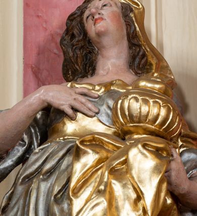 Zdjęcie nr 8: Figura drążona z tyłu, ustawiona na nieregularnej podstawie. Święta zwrócona w trzech czwartych w prawo, w kontrapoście, odchylona łukowato, z głową skierowaną w górę. Prawą dłoń kładzie na piersi, lewą ręką podtrzymuje przy boku płaszcz i puszkę. Twarz ma pociągłą, o podkreślonej brodzie, dużych, głęboko osadzonych, półprzymkniętych oczach, garbatym nosie i małych, pełnych ustach. Włosy długie, gęste, falowane. Ubrana jest w srebrzoną, obfitą i silnie drapowaną suknię o szerokich rękawach, odsłaniającą prawe ramię, przepasaną w talii złoconym gorsetem. Na lewą stronę głowy ma narzucony złocony płaszcz, opadający na lewy bok, silnie drapowany. Polichromia ciała naturalistyczna. 


