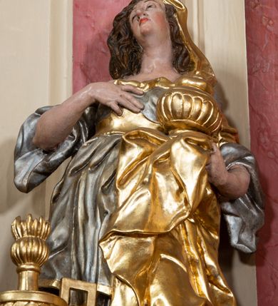 Zdjęcie nr 7: Figura drążona z tyłu, ustawiona na nieregularnej podstawie. Święta zwrócona w trzech czwartych w prawo, w kontrapoście, odchylona łukowato, z głową skierowaną w górę. Prawą dłoń kładzie na piersi, lewą ręką podtrzymuje przy boku płaszcz i puszkę. Twarz ma pociągłą, o podkreślonej brodzie, dużych, głęboko osadzonych, półprzymkniętych oczach, garbatym nosie i małych, pełnych ustach. Włosy długie, gęste, falowane. Ubrana jest w srebrzoną, obfitą i silnie drapowaną suknię o szerokich rękawach, odsłaniającą prawe ramię, przepasaną w talii złoconym gorsetem. Na lewą stronę głowy ma narzucony złocony płaszcz, opadający na lewy bok, silnie drapowany. Polichromia ciała naturalistyczna. 


