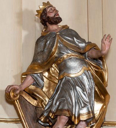 Zdjęcie nr 1: Rzeźba drążona z tyłu, ustawiona na wysokiej, nieregularnej podstawie. Dawid zwrócony w trzech czwartych w lewo, w kontrapoście, łukowato odchylony, z głową skierowaną w górę, lewą rękę wyciąga przed siebie, prawą opiera na złoconej harfie stojącej obok niego. Twarz ma szczupłą, o podkreślonych kościach policzkowych, dużych, migdałowatych oczach, głęboko osadzonych, skierowanych do góry, garbatym nosie i pełnych ustach, z dużą górną wargą. Twarz okolona krótką, gęstą brodą oraz krótkimi, falowanymi, brązowymi włosami. Na głowie ma złoconą koronę otwartą. Ubrany jest w srebrzoną tunikę, sięgającą kolan, obszytą u dołu złoconą frędzlą, w pasie ma srebrzony pancerz, pokryty łuską, złocony przy krawędzi. Na ramiona ma narzucony srebrzony płaszcz, o złoconej podszewce, zapięty pod szyją, a na nogach wysokie, czarne buty, ze złoconą cholewą. U nóg Dawida leżą dwie martwe postacie wrogów ukazanych w popiersiu z hełmami na głowach. Polichromia ciała naturalistyczna. 