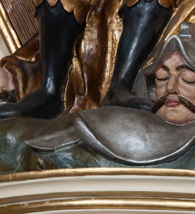 Zdjęcie nr 7: Rzeźba drążona z tyłu, ustawiona na wysokiej, nieregularnej podstawie. Dawid zwrócony w trzech czwartych w lewo, w kontrapoście, łukowato odchylony, z głową skierowaną w górę, lewą rękę wyciąga przed siebie, prawą opiera na złoconej harfie stojącej obok niego. Twarz ma szczupłą, o podkreślonych kościach policzkowych, dużych, migdałowatych oczach, głęboko osadzonych, skierowanych do góry, garbatym nosie i pełnych ustach, z dużą górną wargą. Twarz okolona krótką, gęstą brodą oraz krótkimi, falowanymi, brązowymi włosami. Na głowie ma złoconą koronę otwartą. Ubrany jest w srebrzoną tunikę, sięgającą kolan, obszytą u dołu złoconą frędzlą, w pasie ma srebrzony pancerz, pokryty łuską, złocony przy krawędzi. Na ramiona ma narzucony srebrzony płaszcz, o złoconej podszewce, zapięty pod szyją, a na nogach wysokie, czarne buty, ze złoconą cholewą. U nóg Dawida leżą dwie martwe postacie wrogów ukazanych w popiersiu z hełmami na głowach. Polichromia ciała naturalistyczna. 