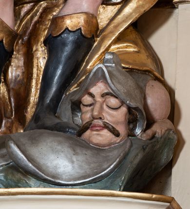 Zdjęcie nr 8: Rzeźba drążona z tyłu, ustawiona na wysokiej, nieregularnej podstawie. Dawid zwrócony w trzech czwartych w lewo, w kontrapoście, łukowato odchylony, z głową skierowaną w górę, lewą rękę wyciąga przed siebie, prawą opiera na złoconej harfie stojącej obok niego. Twarz ma szczupłą, o podkreślonych kościach policzkowych, dużych, migdałowatych oczach, głęboko osadzonych, skierowanych do góry, garbatym nosie i pełnych ustach, z dużą górną wargą. Twarz okolona krótką, gęstą brodą oraz krótkimi, falowanymi, brązowymi włosami. Na głowie ma złoconą koronę otwartą. Ubrany jest w srebrzoną tunikę, sięgającą kolan, obszytą u dołu złoconą frędzlą, w pasie ma srebrzony pancerz, pokryty łuską, złocony przy krawędzi. Na ramiona ma narzucony srebrzony płaszcz, o złoconej podszewce, zapięty pod szyją, a na nogach wysokie, czarne buty, ze złoconą cholewą. U nóg Dawida leżą dwie martwe postacie wrogów ukazanych w popiersiu z hełmami na głowach. Polichromia ciała naturalistyczna. 