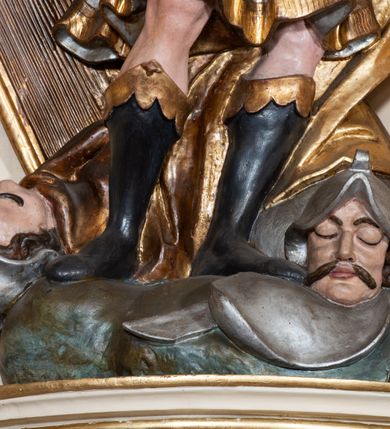 Zdjęcie nr 6: Rzeźba drążona z tyłu, ustawiona na wysokiej, nieregularnej podstawie. Dawid zwrócony w trzech czwartych w lewo, w kontrapoście, łukowato odchylony, z głową skierowaną w górę, lewą rękę wyciąga przed siebie, prawą opiera na złoconej harfie stojącej obok niego. Twarz ma szczupłą, o podkreślonych kościach policzkowych, dużych, migdałowatych oczach, głęboko osadzonych, skierowanych do góry, garbatym nosie i pełnych ustach, z dużą górną wargą. Twarz okolona krótką, gęstą brodą oraz krótkimi, falowanymi, brązowymi włosami. Na głowie ma złoconą koronę otwartą. Ubrany jest w srebrzoną tunikę, sięgającą kolan, obszytą u dołu złoconą frędzlą, w pasie ma srebrzony pancerz, pokryty łuską, złocony przy krawędzi. Na ramiona ma narzucony srebrzony płaszcz, o złoconej podszewce, zapięty pod szyją, a na nogach wysokie, czarne buty, ze złoconą cholewą. U nóg Dawida leżą dwie martwe postacie wrogów ukazanych w popiersiu z hełmami na głowach. Polichromia ciała naturalistyczna. 