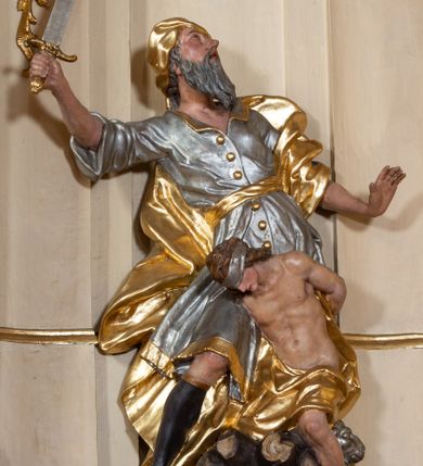 Zdjęcie nr 1: Grupa rzeźbiarska Ofiary Izaaka drążona z tyłu, ustawiona na nieregularnej podstawie. Na stosie porąbanego drewna klęczy Izaak, za nim zaś stoi Abraham zamachujący się na niego mieczem. Abraham w pozycji stojącej, w rozkroku, zwrócony jest w trzech czwartych w lewo, łukowato odchylony w tył, z głową uniesioną do góry. Lewą rękę wyciąga do przodu, prawą unosi wysoko, w dłoni trzyma miecz. Twarz ma szczupłą, o głęboko rzeźbionych oczach skierowanych w górę, z wydrążonym łukiem oczodołu, o ostrym, garbatym nosie oraz pełnych, rozchylonych ustach z dużą górną wargą. Twarz okolona jest siwą, długą, falowaną brodą i krótkimi włosami. Ubrany jest w srebrzoną tunikę z kołnierzykiem, zapinaną na złocone guzy, przepasaną złoconą szarfą, sięgającą kolan, przy dolnej krawędzi dekorowaną złoconą frędzlą. Na lewe ramię ma narzucony złocony płaszcz, opadający na jego prawy bok. Na głowie ma złocony turban, na nogach wysokie brązowe buty ze złoconym brzegiem. Izaak ukazany jako klęczący na prawe kolano na stosie drewna, pochylony, z rękami związanymi za plecami. Twarz ma szczupłą, o garbatym nosie i pełnych ustach, oczy przesłonięte srebrzoną opaską. Włosy ma falowane, opadające na kark. Przesłonięty jest w partii bioder i nóg złoconym płaszczem. Tors silnie umięśniony. Polichromia ciała naturalistyczna.