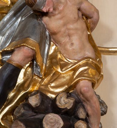 Zdjęcie nr 5: Grupa rzeźbiarska Ofiary Izaaka drążona z tyłu, ustawiona na nieregularnej podstawie. Na stosie porąbanego drewna klęczy Izaak, za nim zaś stoi Abraham zamachujący się na niego mieczem. Abraham w pozycji stojącej, w rozkroku, zwrócony jest w trzech czwartych w lewo, łukowato odchylony w tył, z głową uniesioną do góry. Lewą rękę wyciąga do przodu, prawą unosi wysoko, w dłoni trzyma miecz. Twarz ma szczupłą, o głęboko rzeźbionych oczach skierowanych w górę, z wydrążonym łukiem oczodołu, o ostrym, garbatym nosie oraz pełnych, rozchylonych ustach z dużą górną wargą. Twarz okolona jest siwą, długą, falowaną brodą i krótkimi włosami. Ubrany jest w srebrzoną tunikę z kołnierzykiem, zapinaną na złocone guzy, przepasaną złoconą szarfą, sięgającą kolan, przy dolnej krawędzi dekorowaną złoconą frędzlą. Na lewe ramię ma narzucony złocony płaszcz, opadający na jego prawy bok. Na głowie ma złocony turban, na nogach wysokie brązowe buty ze złoconym brzegiem. Izaak ukazany jako klęczący na prawe kolano na stosie drewna, pochylony, z rękami związanymi za plecami. Twarz ma szczupłą, o garbatym nosie i pełnych ustach, oczy przesłonięte srebrzoną opaską. Włosy ma falowane, opadające na kark. Przesłonięty jest w partii bioder i nóg złoconym płaszczem. Tors silnie umięśniony. Polichromia ciała naturalistyczna.