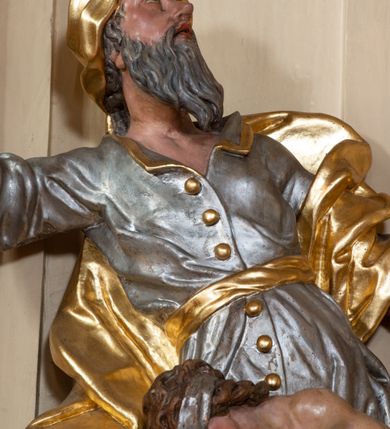 Zdjęcie nr 7: Grupa rzeźbiarska Ofiary Izaaka drążona z tyłu, ustawiona na nieregularnej podstawie. Na stosie porąbanego drewna klęczy Izaak, za nim zaś stoi Abraham zamachujący się na niego mieczem. Abraham w pozycji stojącej, w rozkroku, zwrócony jest w trzech czwartych w lewo, łukowato odchylony w tył, z głową uniesioną do góry. Lewą rękę wyciąga do przodu, prawą unosi wysoko, w dłoni trzyma miecz. Twarz ma szczupłą, o głęboko rzeźbionych oczach skierowanych w górę, z wydrążonym łukiem oczodołu, o ostrym, garbatym nosie oraz pełnych, rozchylonych ustach z dużą górną wargą. Twarz okolona jest siwą, długą, falowaną brodą i krótkimi włosami. Ubrany jest w srebrzoną tunikę z kołnierzykiem, zapinaną na złocone guzy, przepasaną złoconą szarfą, sięgającą kolan, przy dolnej krawędzi dekorowaną złoconą frędzlą. Na lewe ramię ma narzucony złocony płaszcz, opadający na jego prawy bok. Na głowie ma złocony turban, na nogach wysokie brązowe buty ze złoconym brzegiem. Izaak ukazany jako klęczący na prawe kolano na stosie drewna, pochylony, z rękami związanymi za plecami. Twarz ma szczupłą, o garbatym nosie i pełnych ustach, oczy przesłonięte srebrzoną opaską. Włosy ma falowane, opadające na kark. Przesłonięty jest w partii bioder i nóg złoconym płaszczem. Tors silnie umięśniony. Polichromia ciała naturalistyczna.