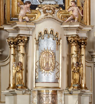 Zdjęcie nr 1: Tabernakulum w formie drewnianej, prostopadłościennej skrzynki, obitej blachą, z wypukłym frontem. Drzwiczki w kształcie stojącego prostokąta, w ramie, dekorowane przedstawieniem kielicha z hostią w glorii, ornamentem roślinnym i uskrzydloną główką anielską. Tron wystawienia ustawiony na dwustrefowym cokole, w którego partii zamontowane jest tabernakulum. Przyjmuje formę drewnianej, prostopadłościennej skrzynki z obrotową niszą, flankowaną dwiema parami kolumienek o kanelowanych trzonach, podtrzymujących fragmenty belkowania, nad niszą przyjmującego kształt falistego gzymsu, na którym znajduje się cokół dekorowany ornamentem roślinnym, z leżącym na księdze barankiem, flankowany dwoma aniołkami, Otwór niszy w formie stojącego prostokąta zamkniętego łukiem półkolistym zdobionym lambrekinem. Przed kolumienkami dwie figurki aniołów trzymających świeczniki.



