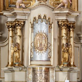 Zdjęcie nr 1: Tabernakulum w formie drewnianej, prostopadłościennej skrzynki, obitej blachą, z wypukłym frontem. Drzwiczki w kształcie stojącego prostokąta, w ramie, dekorowane przedstawieniem kielicha z hostią w glorii, ornamentem roślinnym i uskrzydloną główką anielską. Tron wystawienia ustawiony na dwustrefowym cokole, w którego partii zamontowane jest tabernakulum. Przyjmuje formę drewnianej, prostopadłościennej skrzynki z obrotową niszą, flankowaną dwiema parami kolumienek o kanelowanych trzonach, podtrzymujących fragmenty belkowania, nad niszą przyjmującego kształt falistego gzymsu, na którym znajduje się cokół dekorowany ornamentem roślinnym, z leżącym na księdze barankiem, flankowany dwoma aniołkami, Otwór niszy w formie stojącego prostokąta zamkniętego łukiem półkolistym zdobionym lambrekinem. Przed kolumienkami dwie figurki aniołów trzymających świeczniki.



