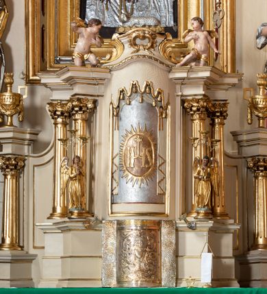 Zdjęcie nr 2: Tabernakulum w formie drewnianej, prostopadłościennej skrzynki, obitej blachą, z wypukłym frontem. Drzwiczki w kształcie stojącego prostokąta, w ramie, dekorowane przedstawieniem kielicha z hostią w glorii, ornamentem roślinnym i uskrzydloną główką anielską. Tron wystawienia ustawiony na dwustrefowym cokole, w którego partii zamontowane jest tabernakulum. Przyjmuje formę drewnianej, prostopadłościennej skrzynki z obrotową niszą, flankowaną dwiema parami kolumienek o kanelowanych trzonach, podtrzymujących fragmenty belkowania, nad niszą przyjmującego kształt falistego gzymsu, na którym znajduje się cokół dekorowany ornamentem roślinnym, z leżącym na księdze barankiem, flankowany dwoma aniołkami, Otwór niszy w formie stojącego prostokąta zamkniętego łukiem półkolistym zdobionym lambrekinem. Przed kolumienkami dwie figurki aniołów trzymających świeczniki.



