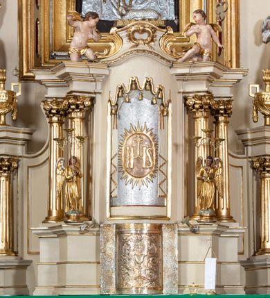 Zdjęcie nr 3: Tabernakulum w formie drewnianej, prostopadłościennej skrzynki, obitej blachą, z wypukłym frontem. Drzwiczki w kształcie stojącego prostokąta, w ramie, dekorowane przedstawieniem kielicha z hostią w glorii, ornamentem roślinnym i uskrzydloną główką anielską. Tron wystawienia ustawiony na dwustrefowym cokole, w którego partii zamontowane jest tabernakulum. Przyjmuje formę drewnianej, prostopadłościennej skrzynki z obrotową niszą, flankowaną dwiema parami kolumienek o kanelowanych trzonach, podtrzymujących fragmenty belkowania, nad niszą przyjmującego kształt falistego gzymsu, na którym znajduje się cokół dekorowany ornamentem roślinnym, z leżącym na księdze barankiem, flankowany dwoma aniołkami, Otwór niszy w formie stojącego prostokąta zamkniętego łukiem półkolistym zdobionym lambrekinem. Przed kolumienkami dwie figurki aniołów trzymających świeczniki.



