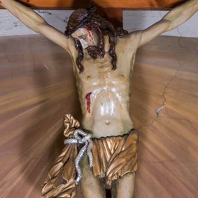 Zdjęcie nr 1: Grupę Ukrzyżowania, usytuowaną w obrębie ściany tęczowej, stanowi sylwetka Jezusa Ukrzyżowanego znajdująca się w górnej partii łuku tęczowego oraz dwie figury asystencyjne, Matki Boskiej i św. Jana Ewangelisty, u jego nasady. Przybitą trzema gwoźdźmi do drewnianego krzyża, pełnoplastyczną figurę Jezusa ukazano w typie Cristo morte. Zwrócona w prawo głowa wsparta jest na ramieniu. Pociągłą twarz charakteryzuje długi, prosty nos oraz krótki, ciemnobrązowy, dwudzielny zarost. Falowane pukle długich włosów opadają na plecy i klatkę piersiową. Ramiona ukształtowane są w głębokim zwisie w kształcie litery „U”. Sylwetkę Jezusa cechuje wydatna muskulatura ciała i sino-blady odcień karnacji. Anatomię torsu podkreśla wyrazista linia żeber. Biodra osłania przewiązane srebrzonym sznurem z prawej strony krótkie, złocone perizonium, z długim zwisem na prawym boku. Lekko ugięte w kolanach nogi przybite są jednym gwoździem. Głowę Jezusa wieńczy korona cierniowa, w prawym boku widoczna jest brocząca krwią rana. Na skroniach, nadgarstkach i stopach są drobne strugi krwi. W górnej partii krzyża biały titulus w formie zwoju, ze złoconym napisem „INRI”. Pod stopami Jezusa, u nasady krzyża jest para uskrzydlonych główek anielskich. Figury asystencyjne znajdują się u nasady łuku tęczowego, ustawione na prostopadłościennych cokołach. Charakteryzują je krępe proporcje sylwetek oraz sumaryczny modelunek fizjonomii i tkanin. Po prawej stronie znajduje się całopostaciowa figura św. Jana Ewangelisty spoglądającego ku górze, ubranego w srebrzoną suknię i jasnoczerwony płaszcz, z prawą ręką wyciągniętą w bok, lewą opuszczoną wzdłuż ciała. Po lewej jest całopostaciowa figura Matki Boskiej także spoglądającej ku górze, o rękach złożonych na piersiach. Ubrana jest w jasnoczerwoną suknię i błękitny, osłaniający głowę płaszcz. 