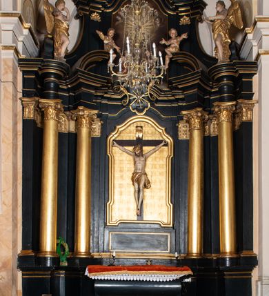 Zdjęcie nr 20: Na dużym krzyżu z ciemnego drewna zawieszono polichromowaną figurę martwego Chrystusa o szeroko rozpostartych ramionach. Ciało Zbawiciela do krzyża jest przymocowane za pomocą trzech gwoździ. On sam został ukazany jako martwy z zamkniętymi oczami i nieznacznie słaniającą się głową. Jego postać jest wysmuklona z realistycznie potraktowaną muskulaturą korpusu. Wyraźnie zaznaczono naprężone ścięgna i mięśnie na całym ciele, co ma sprawiać wrażenie jego autentyczności. Mocno również zaznaczono krwawiącą wybroczynę z przebitego prawego boku. Ciało jest wychudzone o zapadłej klatce piersiowej z silnie podkreśloną linią żeber i mostkiem. Prawa stopa założona jest na lewą. Twarz Zbawiciel jest pociągłą z wyrazistym nosem i wąskim, nieznacznie rozchylonymi ustami. Głowę otaczają brązowe włosy spływające puklami na oba ramiona oraz brązowa broda. Chrystus ma na głowie złoconą, cierniową koronę z długimi cierniami, spod której na czoło spływają krople krwi. Biodra Zbawiciela przepasane są złoconym, krótkim perizonium w formie chusty przerzuconej przez podwójny sznur, odsłaniającej lewe udo i układające się w feston z prawej strony. Ponad głową Chrystusa na pionowej belce krzyża zawieszono rozwiany titulus z napisem "IN / RI".