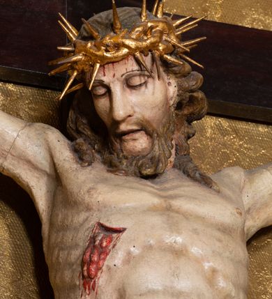 Zdjęcie nr 16: Na dużym krzyżu z ciemnego drewna zawieszono polichromowaną figurę martwego Chrystusa o szeroko rozpostartych ramionach. Ciało Zbawiciela do krzyża jest przymocowane za pomocą trzech gwoździ. On sam został ukazany jako martwy z zamkniętymi oczami i nieznacznie słaniającą się głową. Jego postać jest wysmuklona z realistycznie potraktowaną muskulaturą korpusu. Wyraźnie zaznaczono naprężone ścięgna i mięśnie na całym ciele, co ma sprawiać wrażenie jego autentyczności. Mocno również zaznaczono krwawiącą wybroczynę z przebitego prawego boku. Ciało jest wychudzone o zapadłej klatce piersiowej z silnie podkreśloną linią żeber i mostkiem. Prawa stopa założona jest na lewą. Twarz Zbawiciel jest pociągłą z wyrazistym nosem i wąskim, nieznacznie rozchylonymi ustami. Głowę otaczają brązowe włosy spływające puklami na oba ramiona oraz brązowa broda. Chrystus ma na głowie złoconą, cierniową koronę z długimi cierniami, spod której na czoło spływają krople krwi. Biodra Zbawiciela przepasane są złoconym, krótkim perizonium w formie chusty przerzuconej przez podwójny sznur, odsłaniającej lewe udo i układające się w feston z prawej strony. Ponad głową Chrystusa na pionowej belce krzyża zawieszono rozwiany titulus z napisem &quot;IN / RI&quot;.